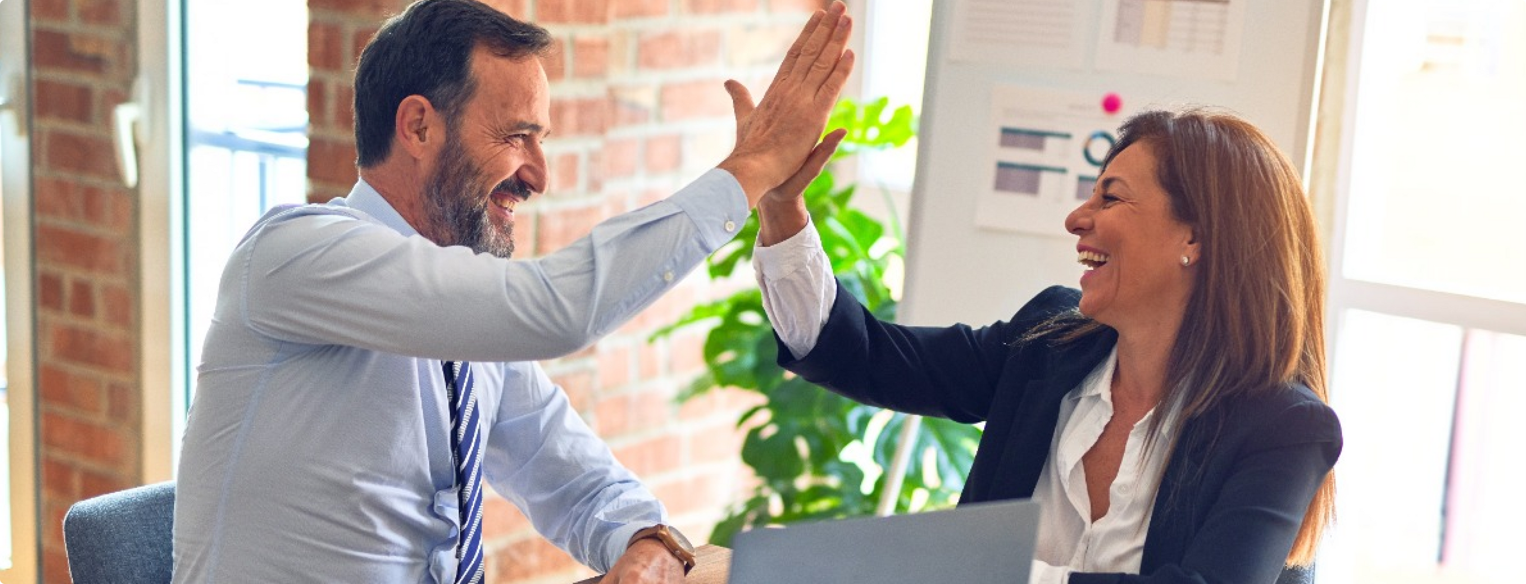 Ein Mann und eine Frau in Business-Kleidung lächeln und geben sich in einem Büro ein High-Five. Auf dem Schreibtisch vor ihnen steht ein geöffneter Laptop, im Hintergrund sind eine Pflanze und einige Diagramme zu sehen.