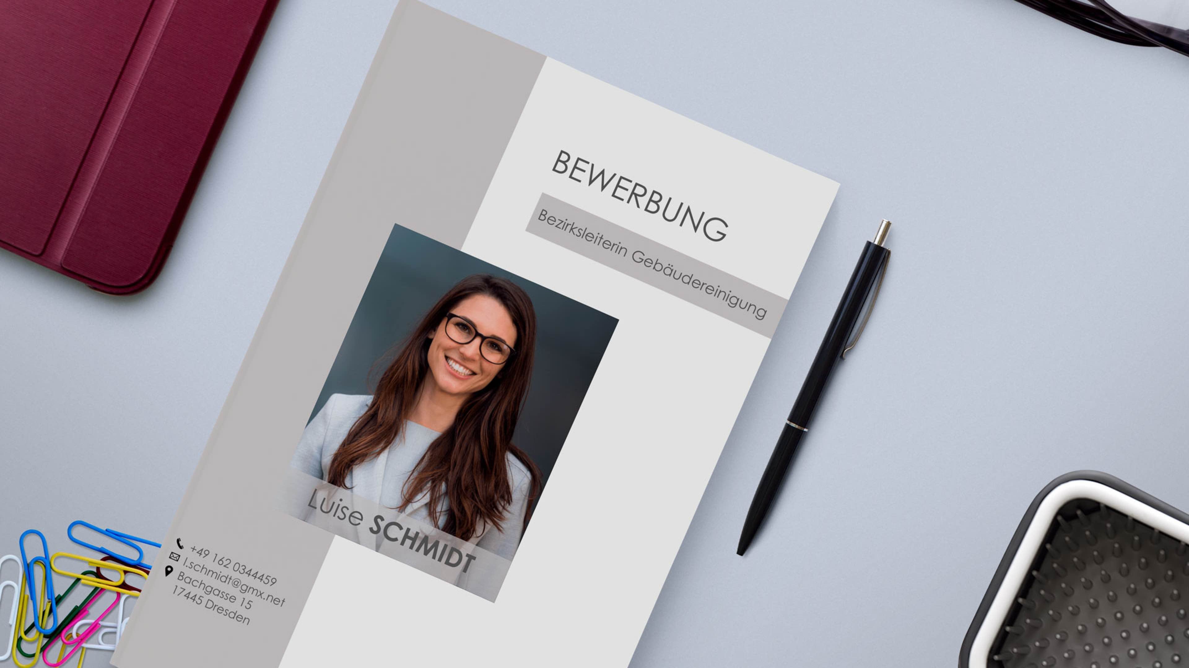 Ein Bewerbungsschreiben für eine Reinigungsfachkraft namens Luise Schmidt mit ihrem Foto. Es liegt auf einem Schreibtisch. Zu sehen sind ein Stift, Büroklammern, ein Ordner und ein Teil einer Tastatur.