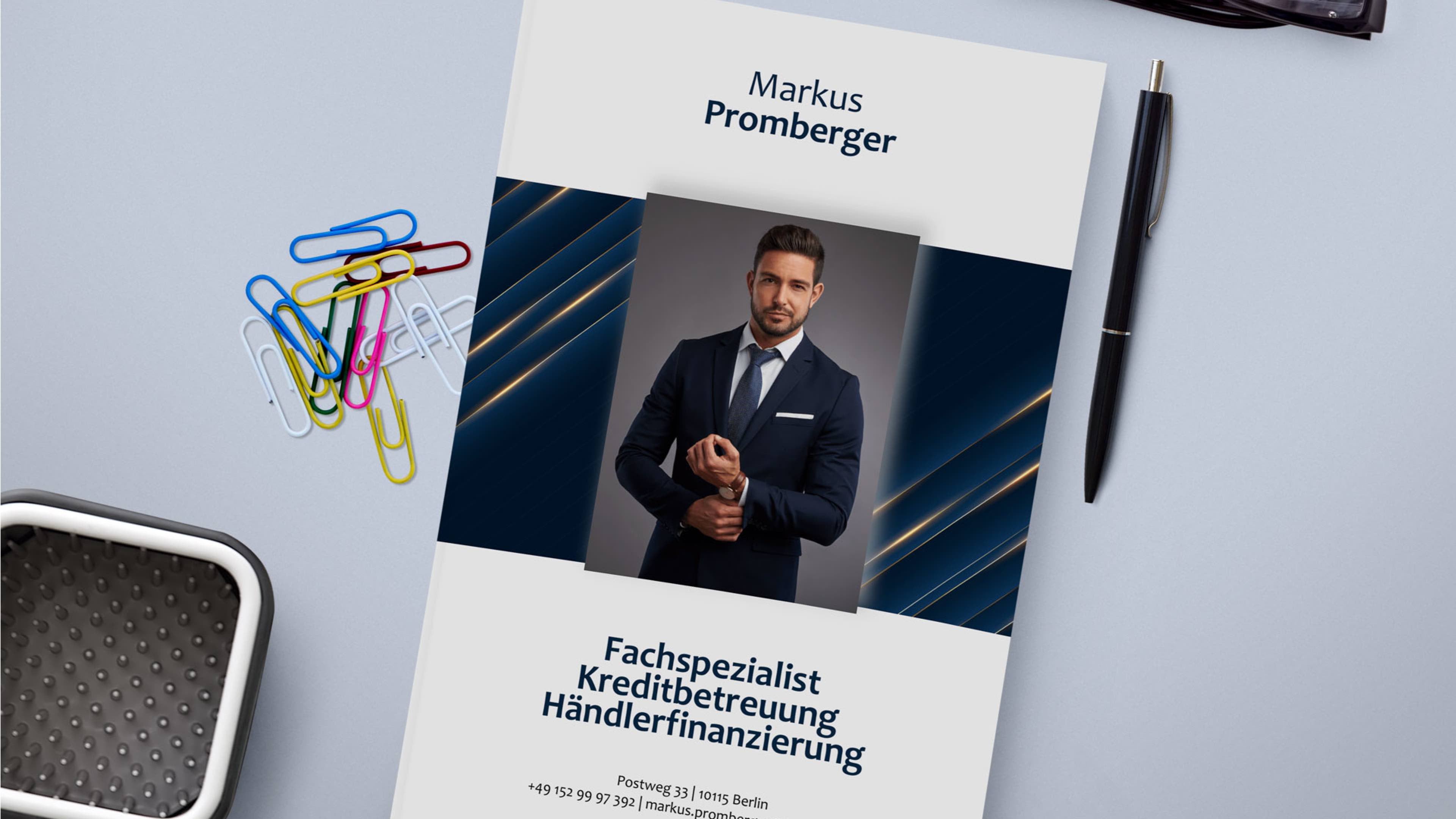 Auf einem Schreibtisch liegt eine Broschüre mit dem Porträt eines Mannes im Anzug. Im Text steht „Markus Promberger“ und „Fachspezialist Kreditbetreuung Händlerfinanzierung“. Verschiedene farbige Büroklammern und ein Stift liegen in der Nähe.