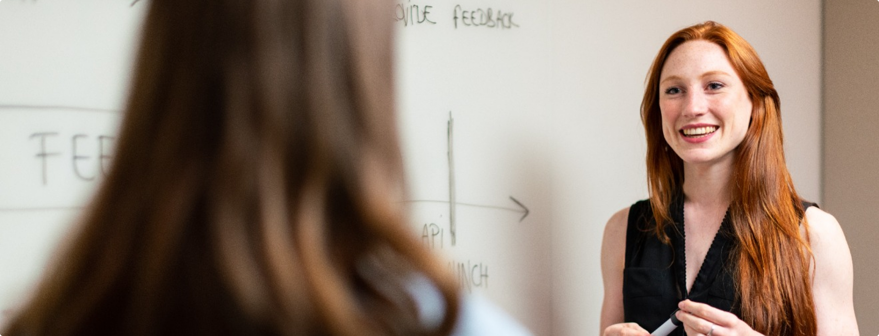 Eine Frau mit langen roten Haaren und einem schwarzen Oberteil lächelt und gestikuliert vor einer Tafel mit Diagrammen. Sie unterhält sich mit einer anderen Person, die der Kamera den Rücken zuwendet.