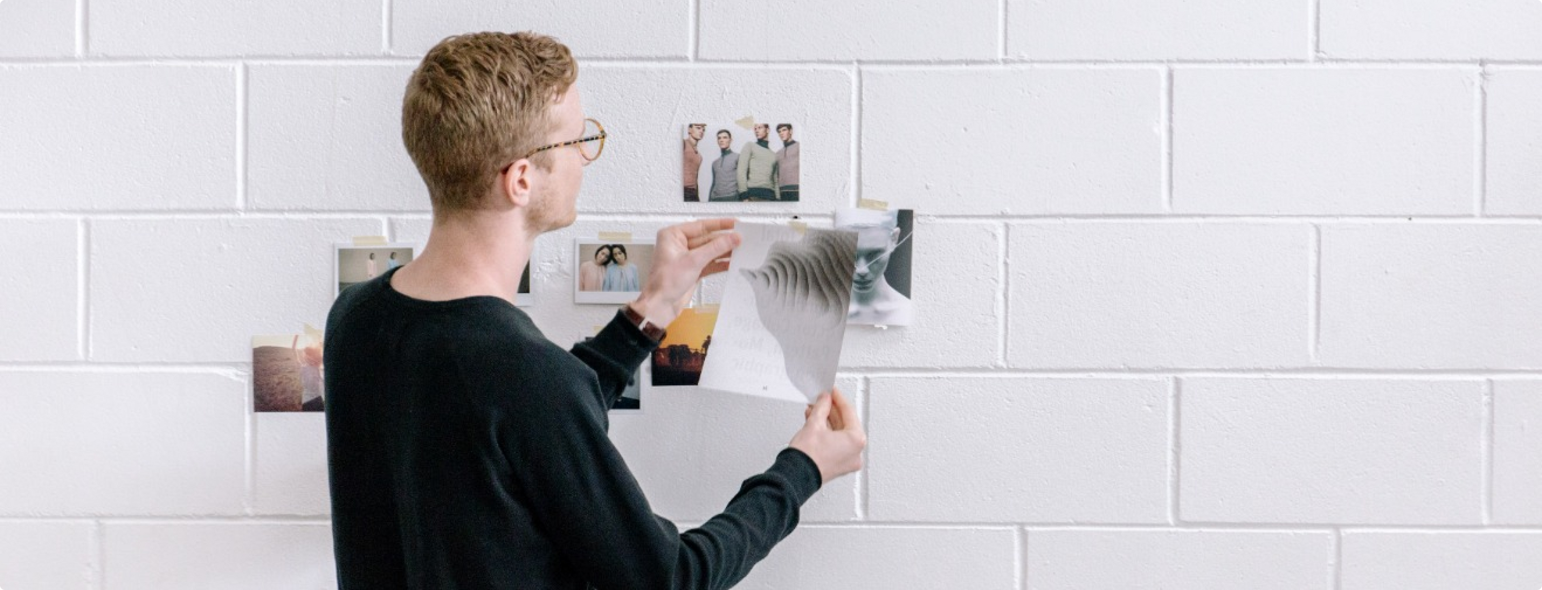 Eine Person mit kurzen Haaren und Brille pinnt ein Dokument an eine weiße Backsteinwand. Mehrere Fotos sind bereits an der Wand befestigt. Die Person trägt einen schwarzen Pullover und scheint konzentriert die Gegenstände zu ordnen.